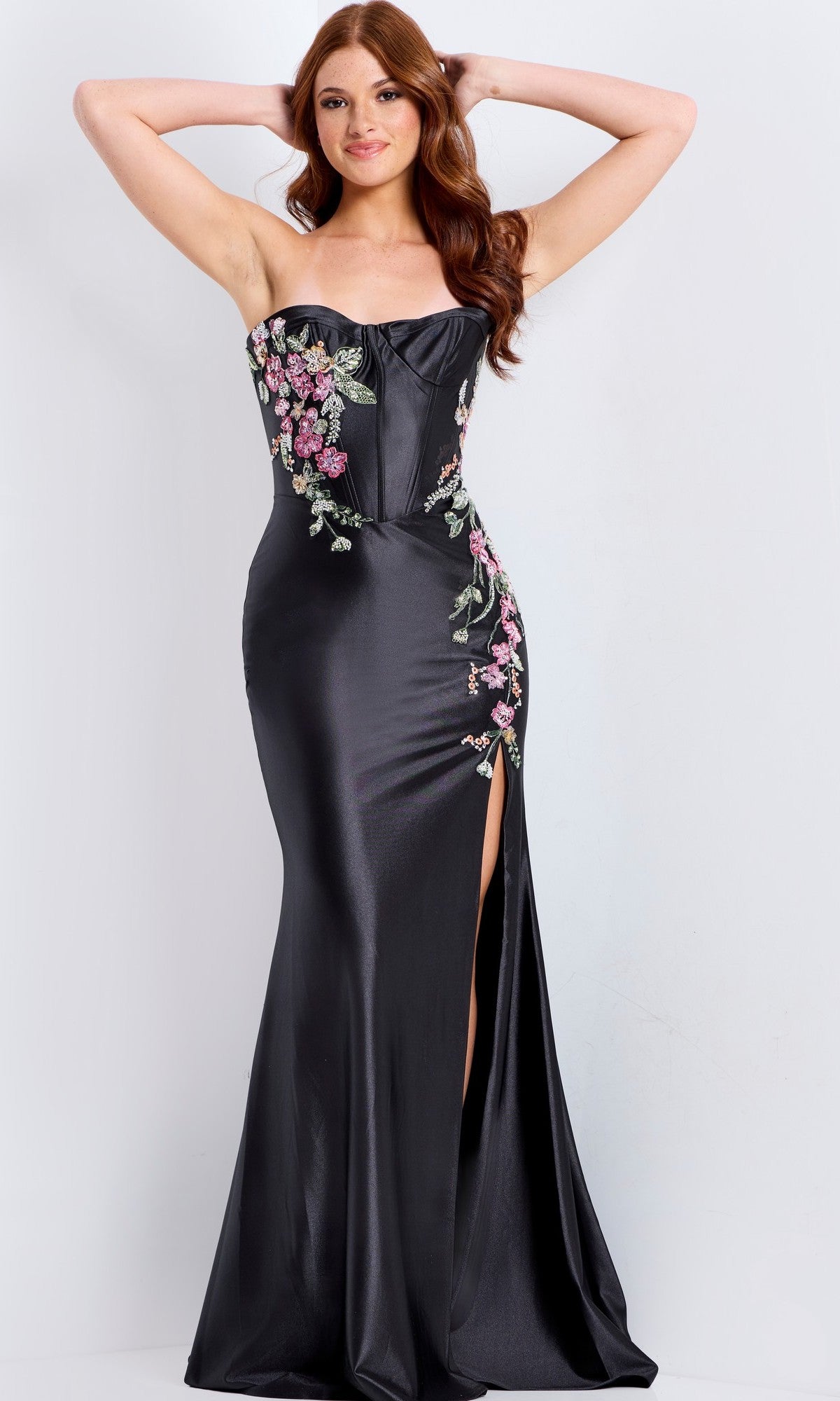 A woman poses with hands behind her head in the Jovani 48371 long prom dress, a black strapless gown featuring floral embroidery, a fitted corset bodice, and a thigh-high slit, set against a plain background.